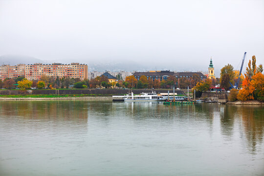 Danube River in Budapest, Hungary, looking towards the Buda side hilly area from the flatter Pest riverfront. Buildings on the bank include a prominent church steeple with boats moored along shore