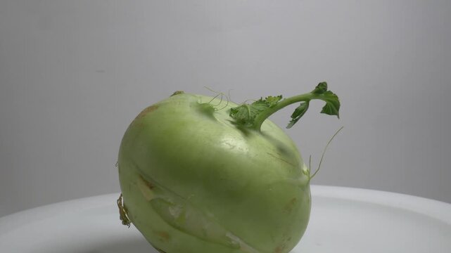 A fresh kohlrabi bulb with a small green leaf rotates slowly on a white plate in a studio setting, showcasing its unique shape and natural skin texture in close-up.