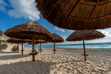 Fototapeta premium Sombrillas de palapa sobre arena blanca frente al mar Caribe con el sol brillando entre las nubes. Escena tropical en la Riviera Maya que evoca vacaciones, descanso y estilo de vida relajado junto al 