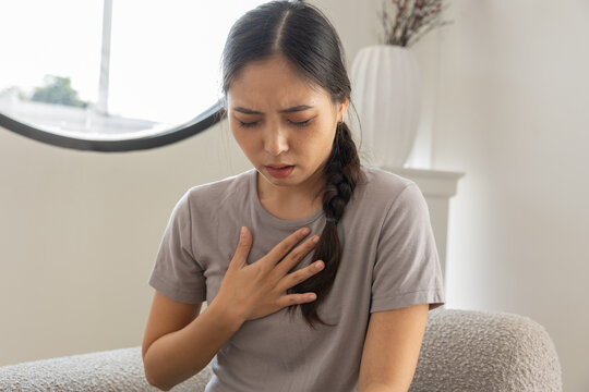 Young distressed Asian woman feeling sharp chest pain breathing hard suffering heart attack or acid reflux symptoms while sitting home alone unwell sick female patient clutching chest having
