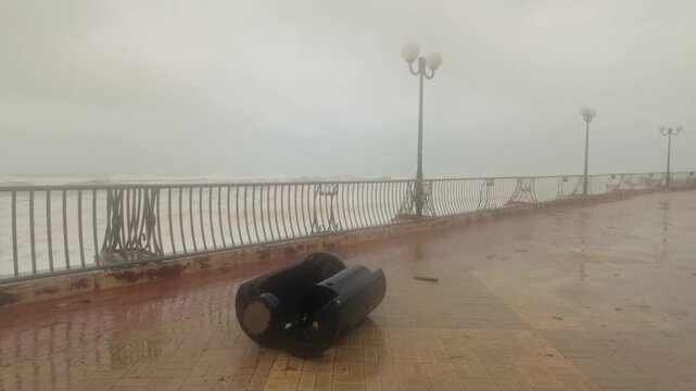 Storm Harry lashes Marsaskala, Malta, as strong winds and heavy rain overturn a garbage bin on the seaside promenade, with rough Mediterranean waves and an overcast winter sky in the background.