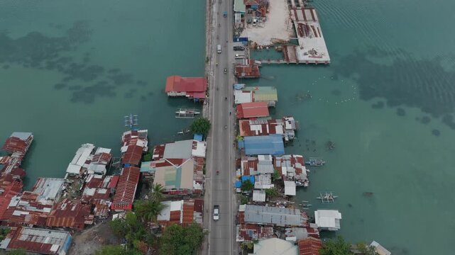 Vehicles move steadily across causeway of Tagbilaran City and Dauis, homes built up on edge in shacks, aerial pullback overview