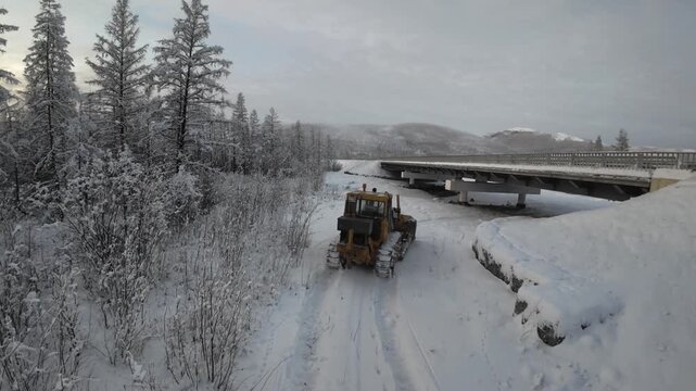 A yellow snowplow clears a snowy road under a bridge in a winter forest, emphasizing outdoor winter maintenance.