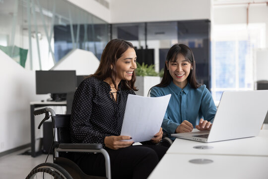 Happy female Middle Eastern office employee with disability consulting young Chinese colleague. Diverse coworkers of inclusive company working together at laptop, typing, smiling