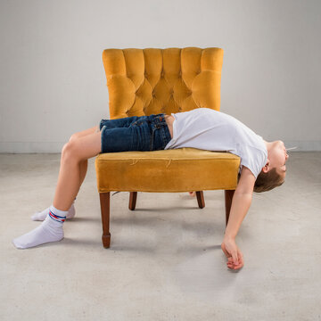 vintage retro young preteen male boy child wearing tee shirt jean shorts socks sitting reclining laying on vintage retro chair in empty space with dramatic lighting with sucker candy