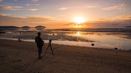 A lone photographer stands on a quiet beach with a tripod, capturing a golden sunrise over shallow tidal waters. The expansive sky, reflections on wet sand, and distant islands create a calm  © Narong Niemhom