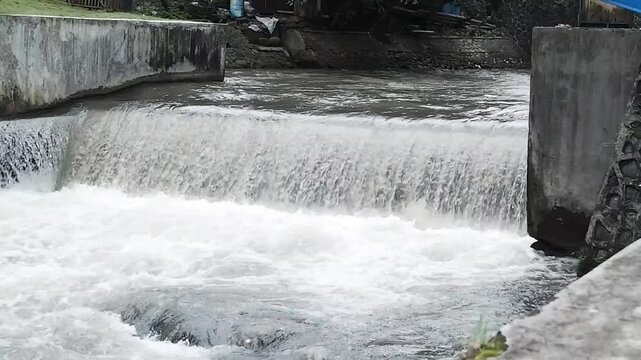 Fast-flowing river water cascades over a small concrete weir, creating white foam and dynamic currents, illustrating water management, natural motion, and rural infrastructure, landscape view. 