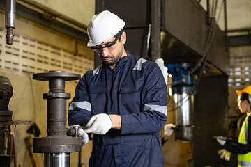 Concentrated young mechanical engineer in hard hat using wrench to repair machine in industrial factory with professional serious emotion while mechanic colleague working