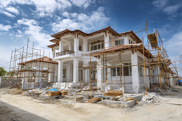 Unfinished residential house under construction with scaffold surrounding building, clear blue sky and scattered clouds above, construction materials on ground