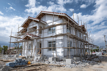 Unfinished residential house under construction with scaffold surrounding white two story building, clear sky and scattered construction materials on ground creating busy worksite atmosphere