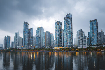 Fototapeta premium Urban skyline with modern skyscraper buildings reflecting on calm water under cloudy sky, creating dramatic and serene cityscape atmosphere at dusk
