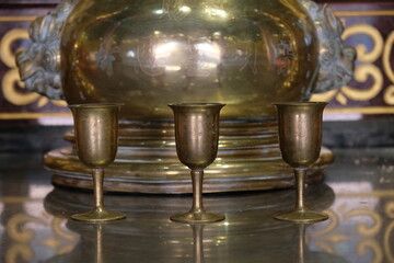 Three antique brass chalices stand before a large ornate golden vessel in a dimly lit sacred space © Thanakrit