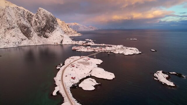 Cinematic 4K drone shot of Hamnoy village at sunset. Iconic red rorbuer cabins against snowy mountain and reine fjord in Lofoten, Norway. Winding E10 road and bridge conecting small islands