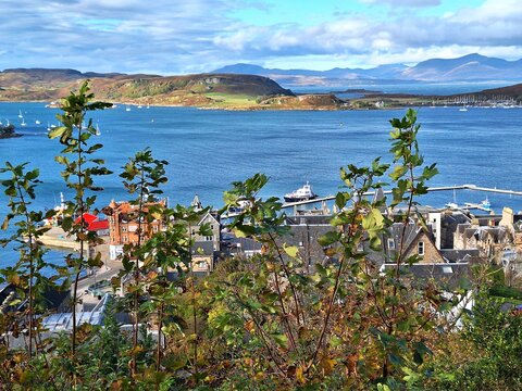 Panoramic view over Oban Bay in Argyll and Bute, Scotland, captures blue waters dotted with boats and yachts, rolling hills in the distance, and traditional stone buildings clustered along shoreline.