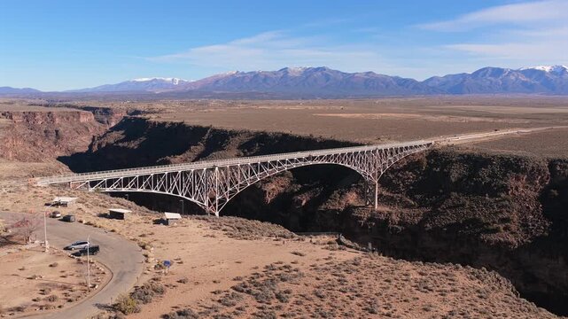 The drone flies forward toward the steel arch Rio Grande Gorge Bridge. The high desert landscape features deep basalt rock walls and the snow-capped Sangre de Cristo Mountains under a blue sky.