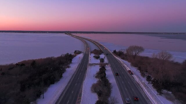An aerial view of the Robert Moses Causeway and the Great South Bay during a cold and colorful sunset. The camera dolly in between the roads as cars drive by in the wintery scene.