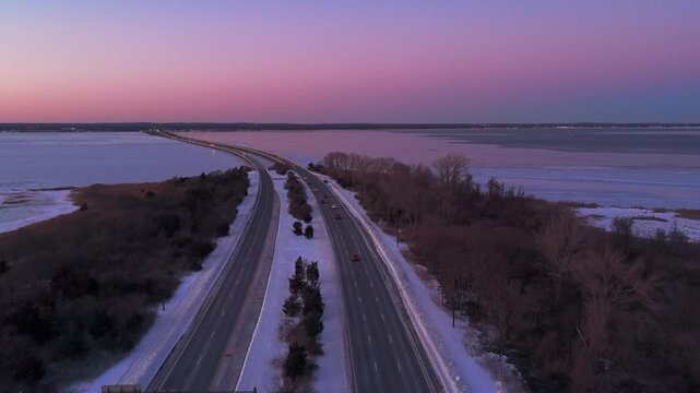 An aerial view of the Robert Moses Causeway and the Great South Bay during a colorful but cold sunset. The camera dolly in between the roads as cars drive by in the wintery scene.