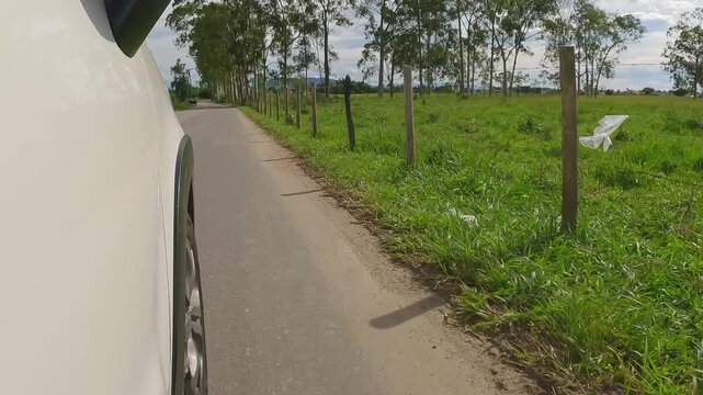 Side point of view shot of a white car driving on a rural dirt road alongside a wooden fence and green pastures. Countryside travel and farm concept.