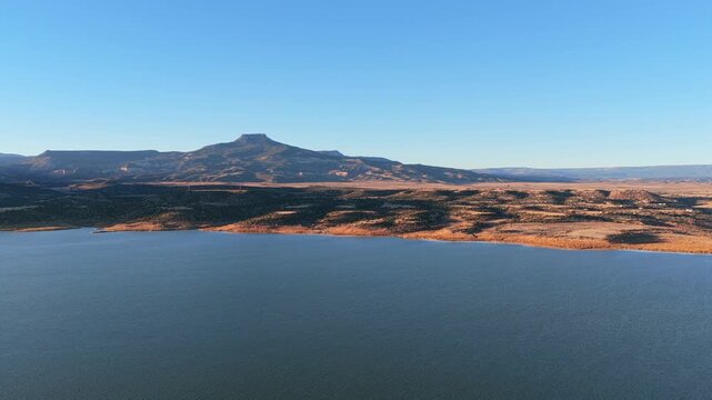 Deep blue water of Abiquiu Lake reservoir borders the arid desert brush of northern New Mexico. The volcanic basalt peak of Cerro Pedernal rises in the distance under a clear and bright blue sky.