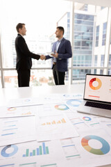 Fototapeta premium Conclusion of successful negotiation, mutual agreement, trust. Close up of laptop view with displaying pie charts data and papers spread out on table, two businessmen shaking hands stand in background