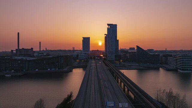 Drone hyperlapse Kulosaarensilta to Kalasatama Helsinki: approach Redi shopping center and towers along It&auml;v&auml;yl&auml; bridge. Urban waterfront scene, sun setting behind high-rises, golden hour atmosphere.