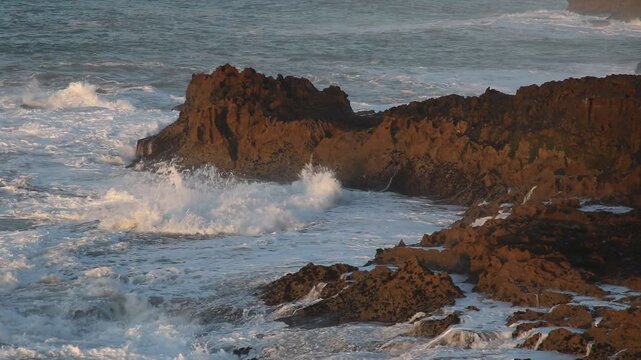 Ocean waves crashing against distinctive red rock formations at Legzira Beach in Sidi Ifni, Morocco, showing white water flow, rugged coastline, natural erosion, and dramatic coastal scenery