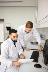 Obraz premium Medical teamwork and collaboration. Confident male doctor clinician healthcare specialist sit at desk in clinic office read paper document with colleague intern discuss diagnosis prepare therapy plan