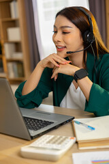 Happy smiling young Asian woman customer service operator with headset working on vertical laptop screen at office desk with calculator while talking to client with confident expression