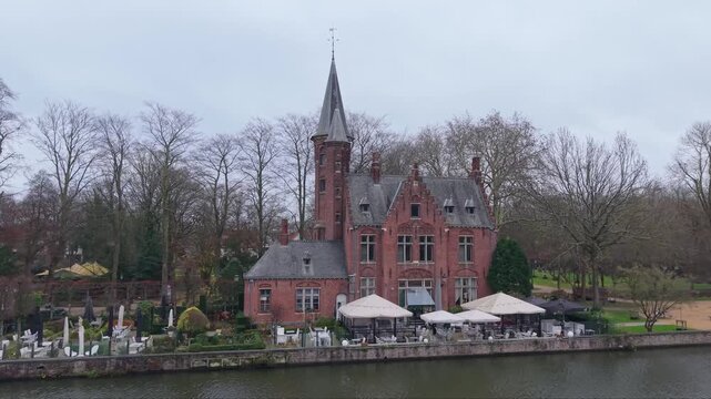 Aerial orbit of the neo-Gothic Kasteel Minnewater (Lake of Love Castle) in Bruges, Belgium. Moody winter atmosphere with historic architecture and bare trees under an overcast sky.