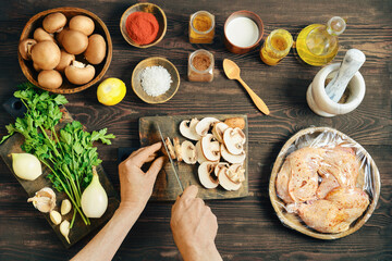 Top view of chef hands slicing fresh mushroom on wooden board with marinated chicken nearby