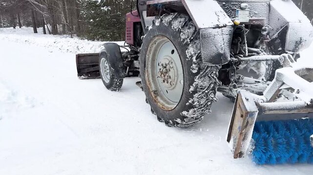 Vintage tractor with blue rotary brush sweeper attachment clearing snow in winter forest road for municipal maintenance and snow removal operations