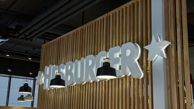 Hesburger fast food restaurant logo sign with white 3D letters and star mounted on wooden slat wall with black pendant lights in modern interior design. 7 February, 2026, Zarasai Lithuania