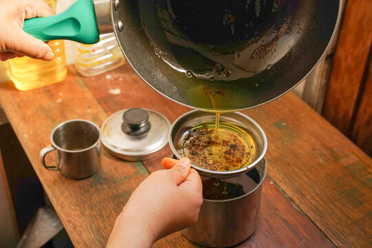 A woman&rsquo;s filtering used cooking oil from non stick pan into a stainless steel container on a wooden kitchen table, for proper disposal and recycling at home