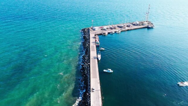 Static drone shot of a Pomorie pier with boats docked on one side and waves breaking against rocks and algae on the other.