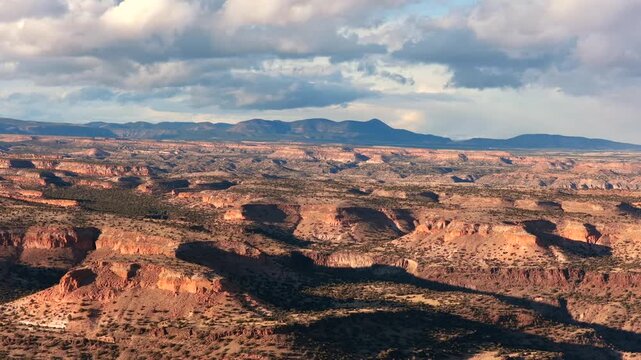 A wide aerial view shows the layered volcanic mesas and dramatic canyon shadows of the Pajarito Plateau. Snow-capped mountains sit on the far horizon under a blue sky with soft white clouds.