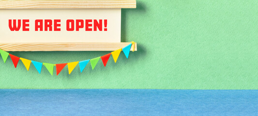 We are open. Invitation greeting card for opening ceremony, start of a project, business launch. wooden sign decorated with garland of flags. green blue background © besjunior