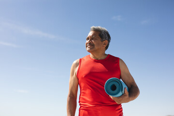 Senior man with rolled up exercise mat looking away at beach against blue sky
