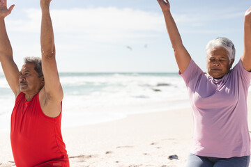 Obraz premium Senior couple exercising together with arms raised at beach against sky on sunny day