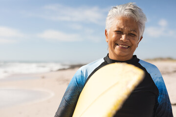 Fototapeta premium Portrait of smiling senior woman carrying surfboard at beach against sky on sunny day