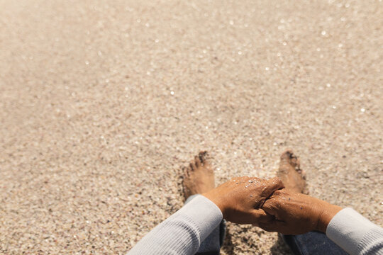 Low section of senior man sitting with hands clasped on sand at beach during sunny day