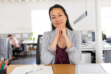 Portrait of confident asian creative businesswoman sitting with hands on chin at desk in office