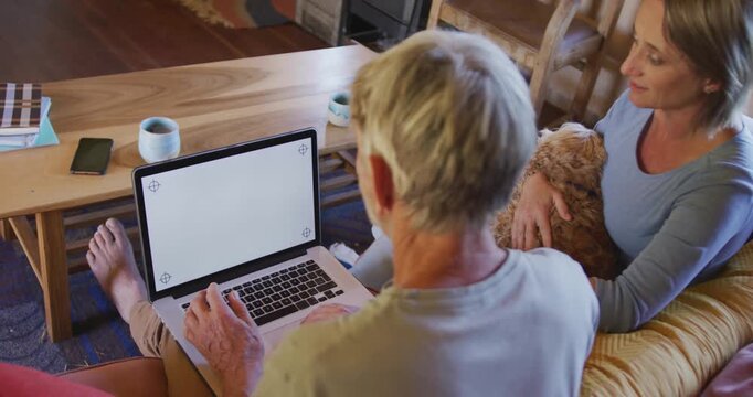 Smiling senior caucasian couple using laptop and sitting with dog in living room