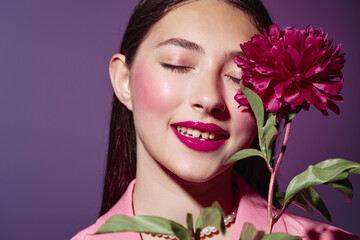 Fototapeta premium Young woman with fashion makeup and vibrant fuchsia lips poses with closed eyes near a matching pink flower. Artistic portrait with bold colors and creative beauty concept.