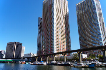 晴れ渡った空の下、運河沿いにそびえ立つ複数のタワーマンションとモノレールの高架線