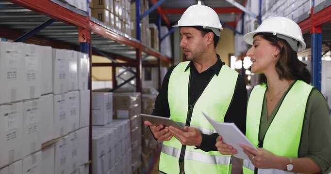 Two Hispanic warehouse employees checking goods for inventory, discussing merchandising, standing at stacked boxes on shelves in storage, talking, holding waybill and tablet
