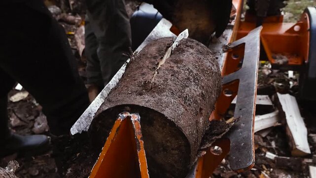 Close view of a compact firewood processor splitting a thick log with a hydraulic ram, while gloved hands work in the background during outdoor wood processing.