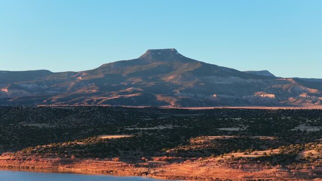 A cinematic drone approach moves closer to a flat-top mountain over a blue lake. Warm sunrise light hits the rugged desert landscape in this high-quality travel background.