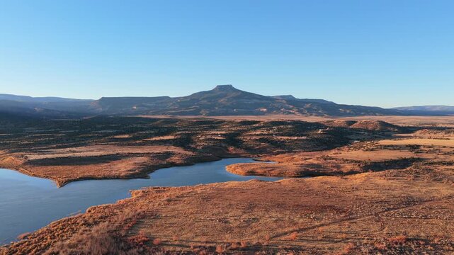 Aerial view captures the blue reservoir water of Abiquiu Lake below the volcanic basalt peak of Cerro Pedernal. Arid brush and sunlit shorelines define the high desert plateau of northern New Mexico.