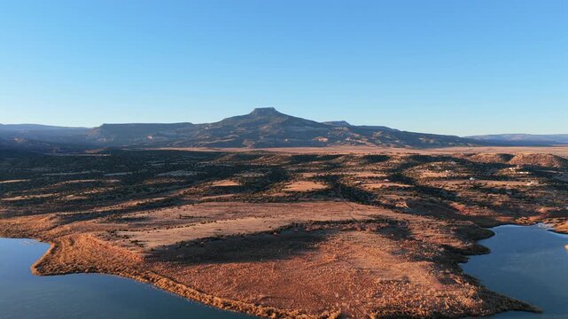Morning sunlight hits the volcanic basalt peak of Cerro Pedernal above Abiquiu Lake. The high desert plateau landscape features arid soil and a rippled blue reservoir in northern New Mexico.