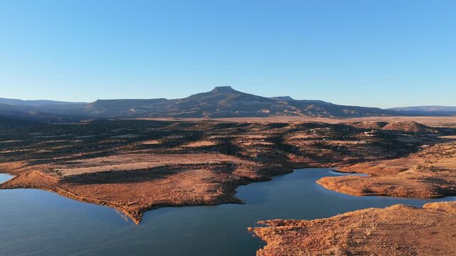 Sunlight hits the arid brush and textured shoreline of Abiquiu Lake reservoir. In the distance, the volcanic basalt cap of Cerro Pedernal mesa sits above the high desert plateau of New Mexico.